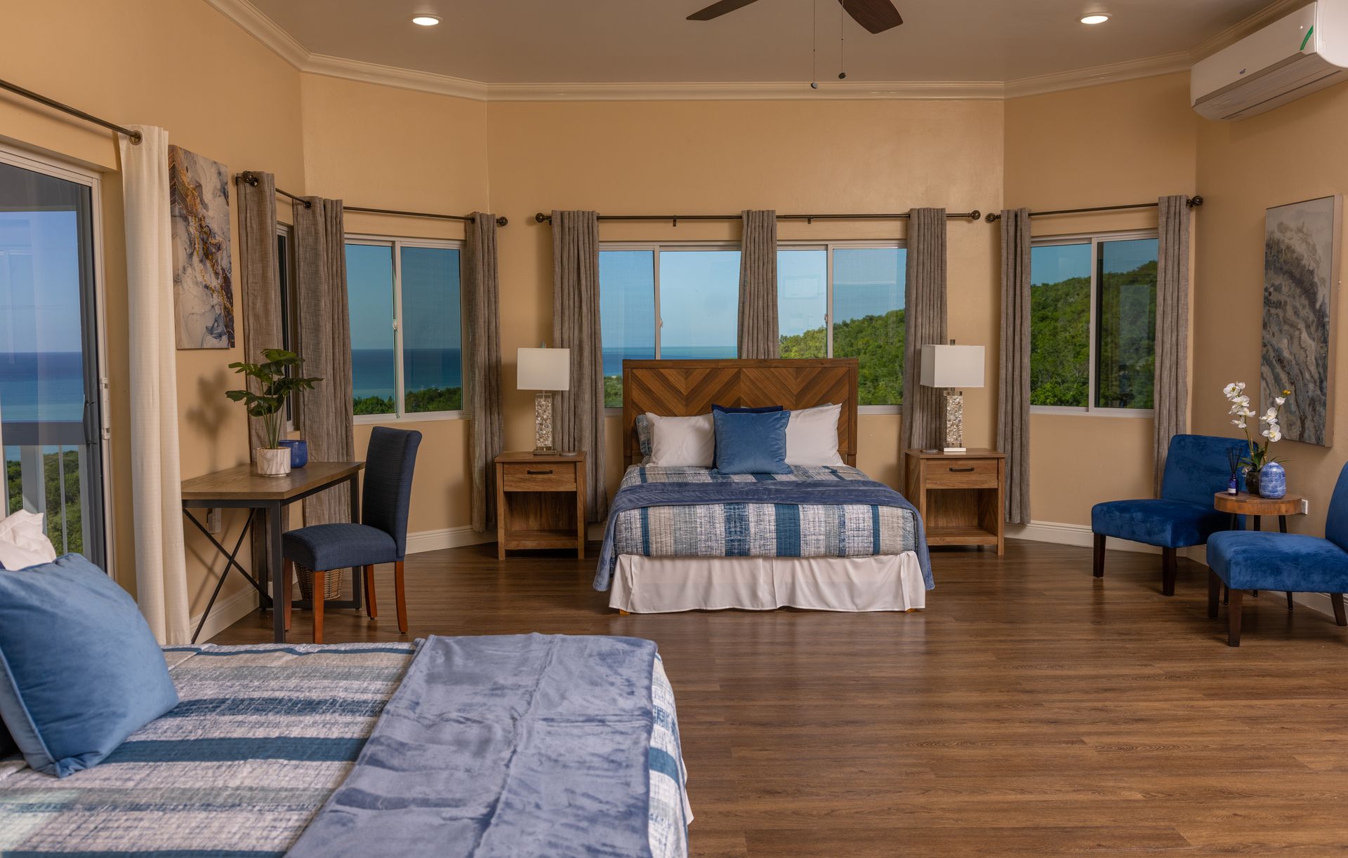 Spacious bedroom with ocean view. Two beds, nightstands, desk, and blue accent chairs.
