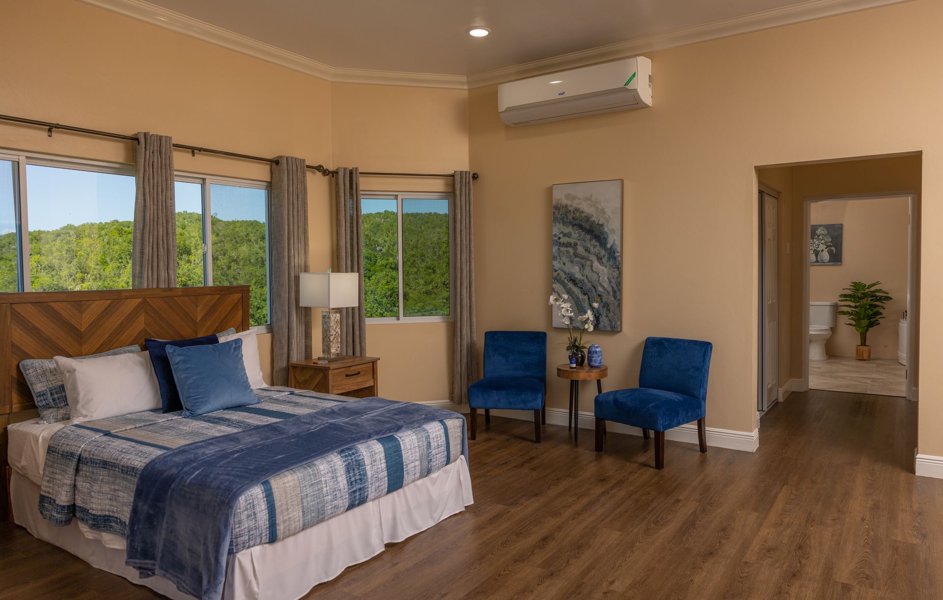 Bedroom with a bed, two blue chairs, and a doorway to a bathroom. Beige walls and hardwood floor.