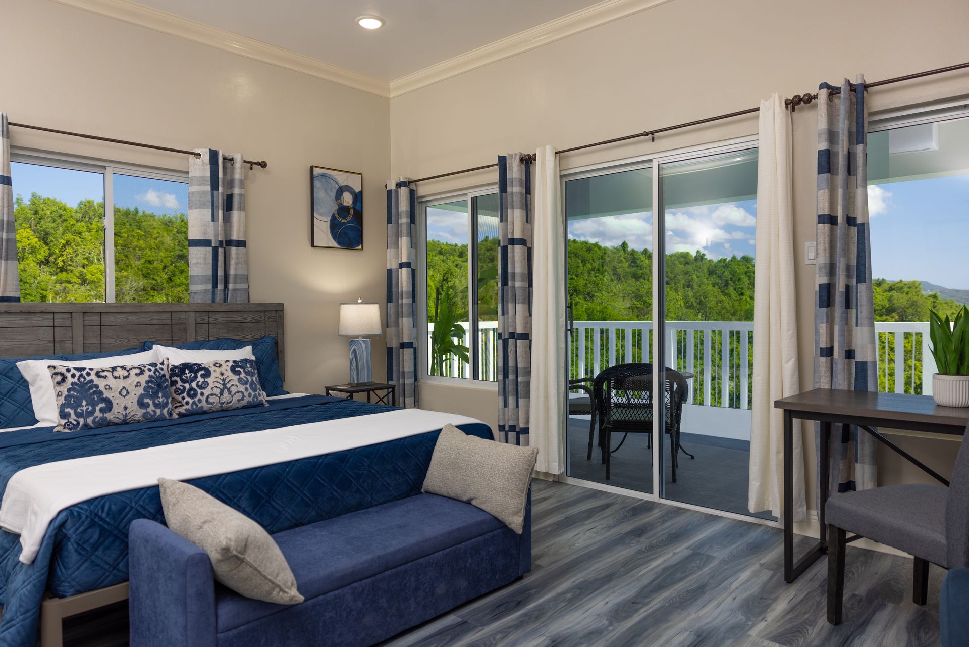 Bedroom with a bed, bench, and sliding doors leading to a balcony with forest view. Blue and gray decor.