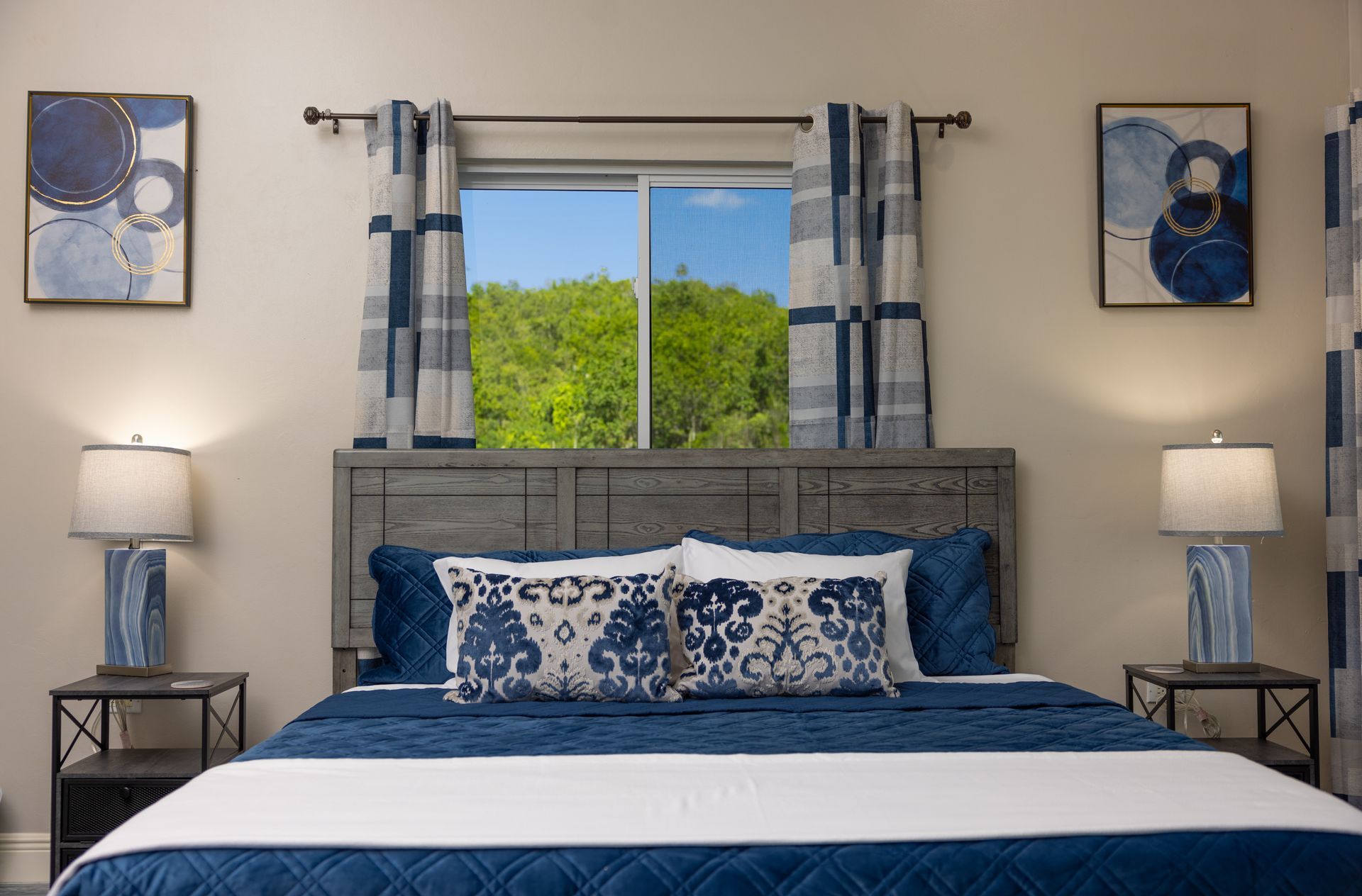 Bedroom with blue and white decor, king bed, artwork, lamps, and a window overlooking greenery.