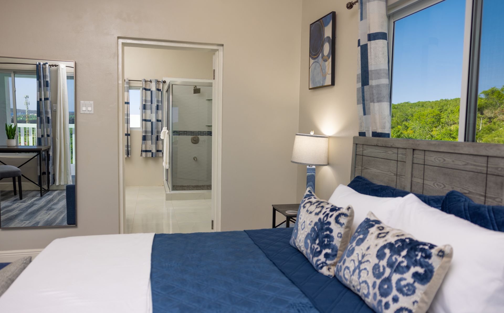 Bedroom with blue and white bedding, wooden headboard, open door to bathroom, and window with trees.