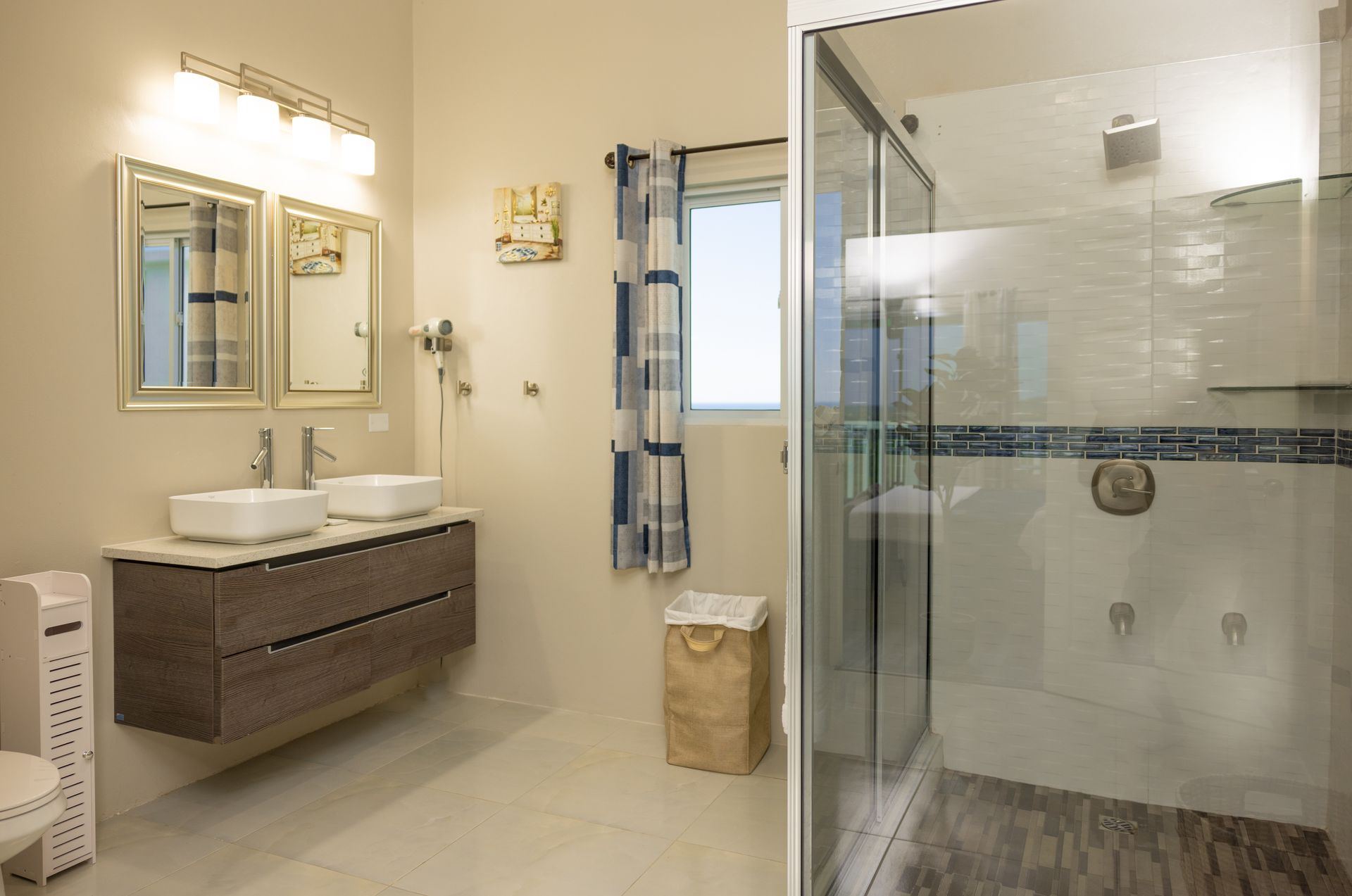 Bathroom with a floating vanity, two white sinks, a glass shower, and a window with blue checkered curtains.