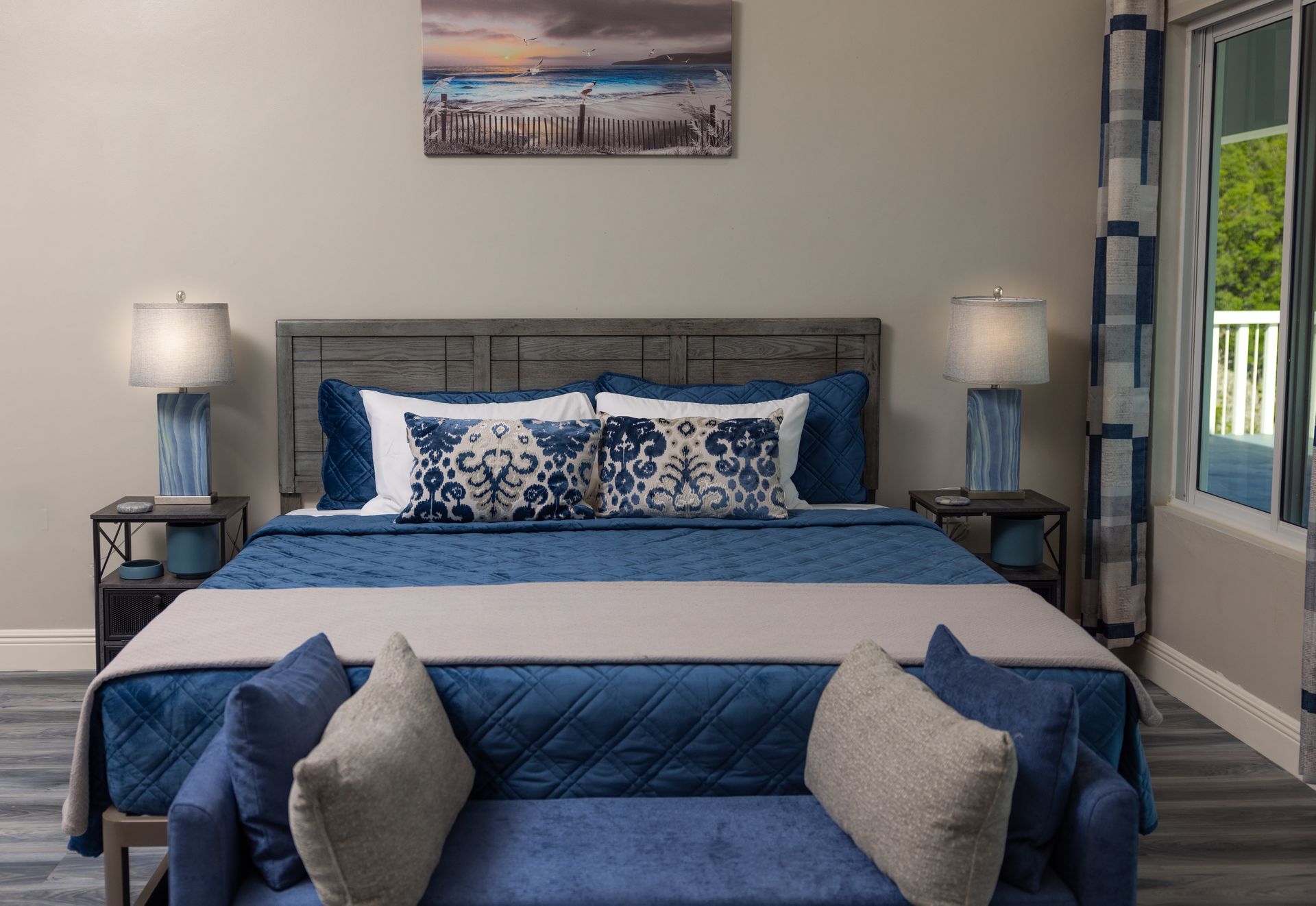 Blue and beige bedroom with king-size bed, nightstands, lamps, and a bench. Coastal themed decor, window to the right.