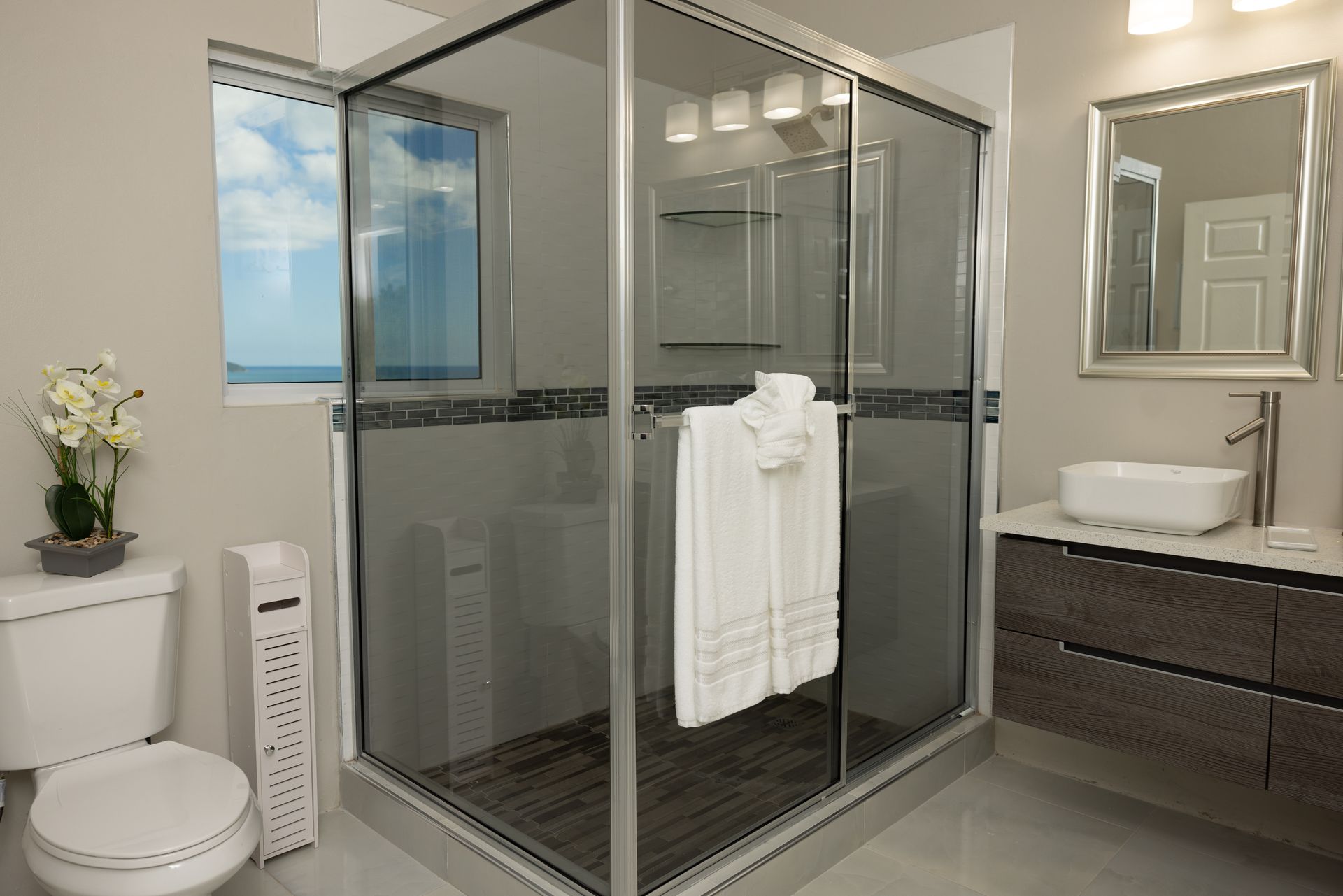Modern bathroom with a glass shower, toilet, sink, and window with a view.