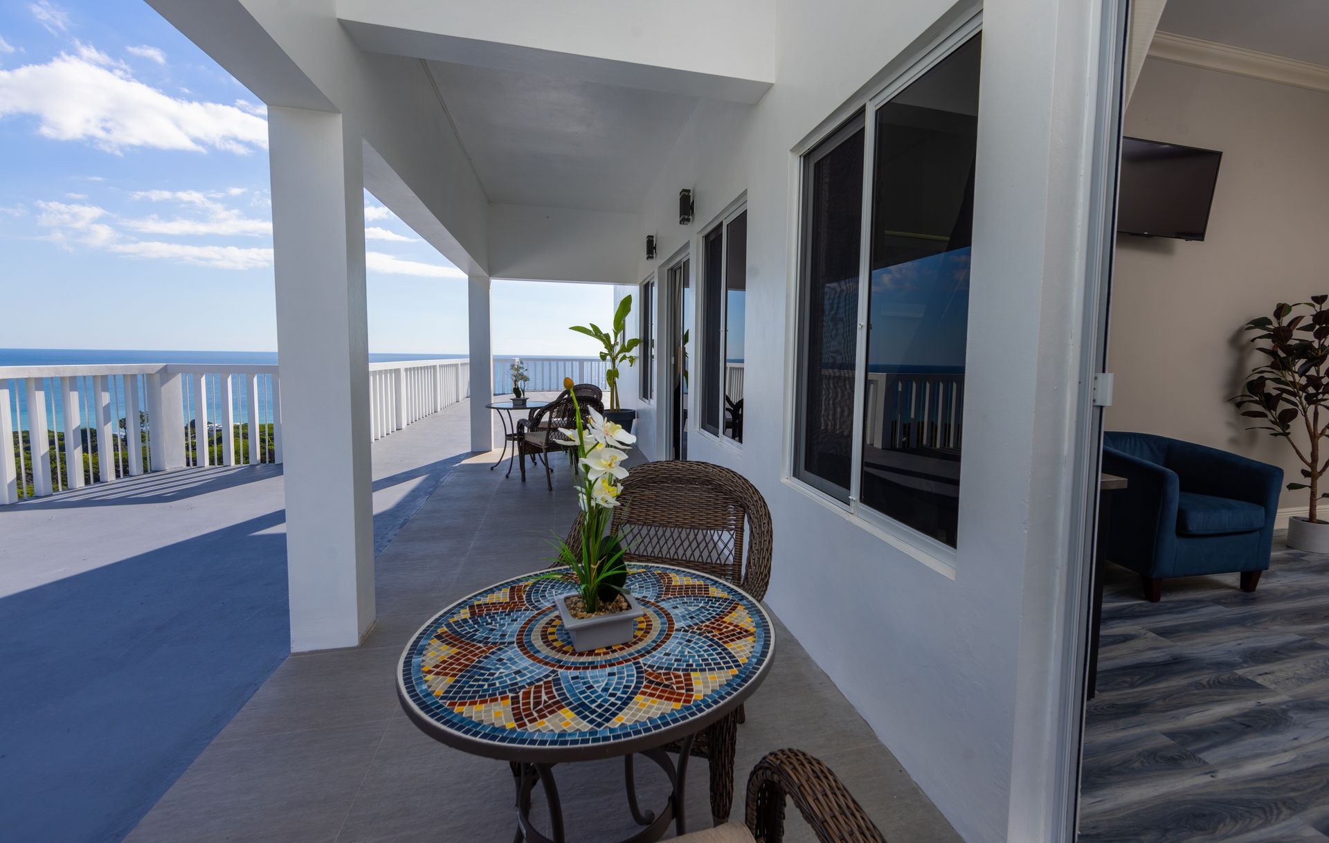Balcony with ocean view, mosaic table, wicker chairs, white pillars, blue sky.