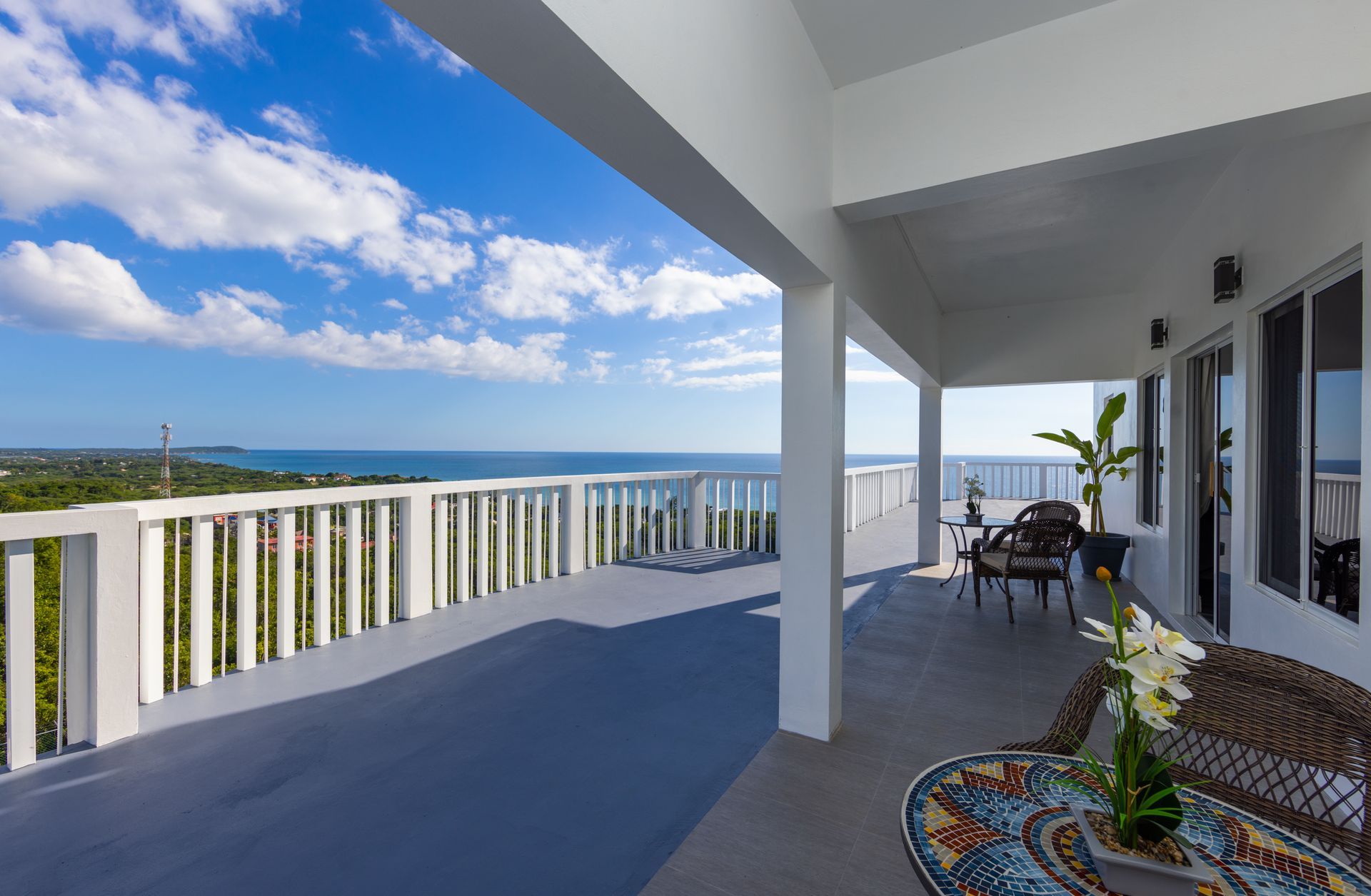 Balcony overlooking ocean; white railing, blue sky, furnished with table and chairs.