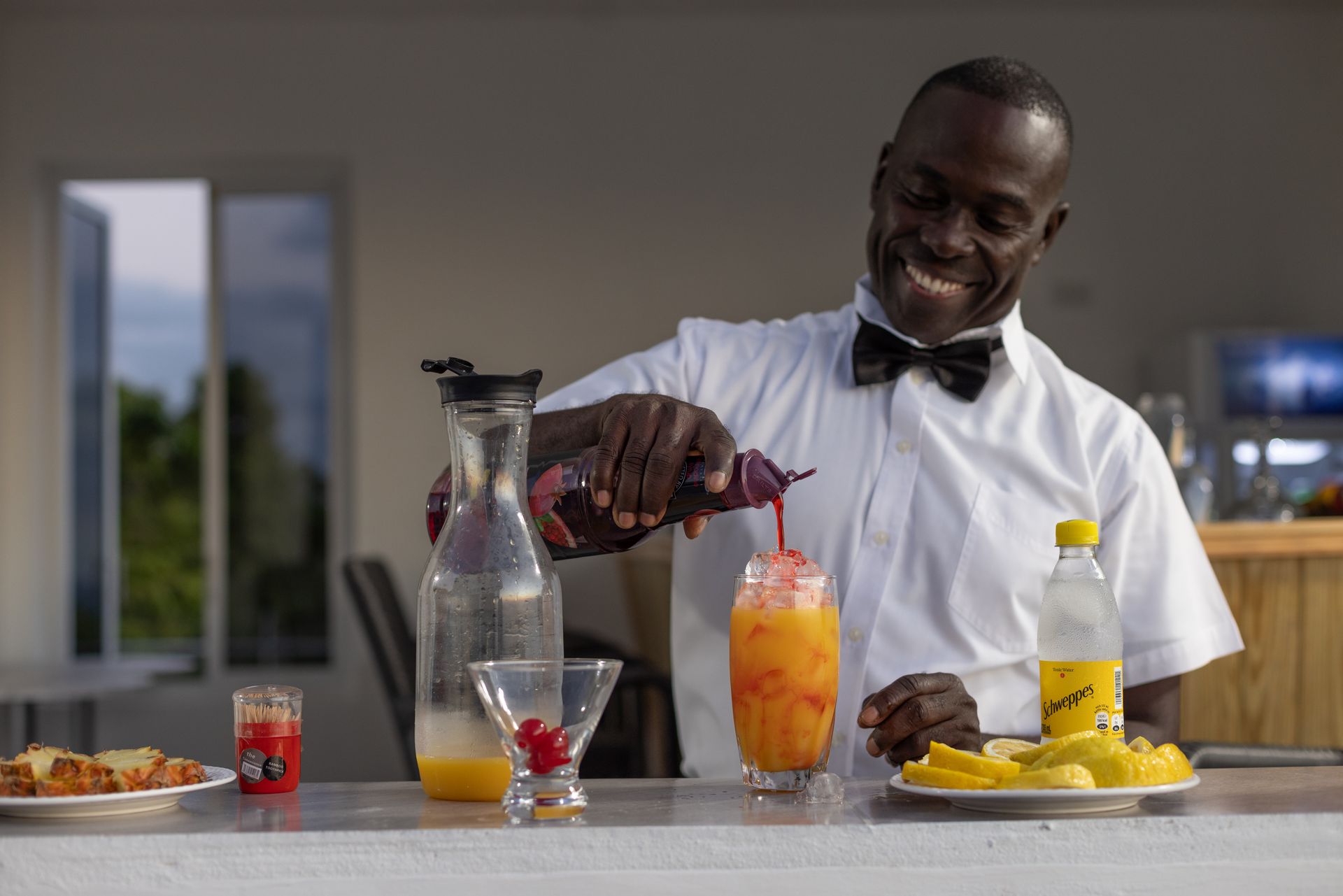 Bartender pouring a colorful drink into a tall glass, smiling. A bar with snacks and other drinks in the background.
