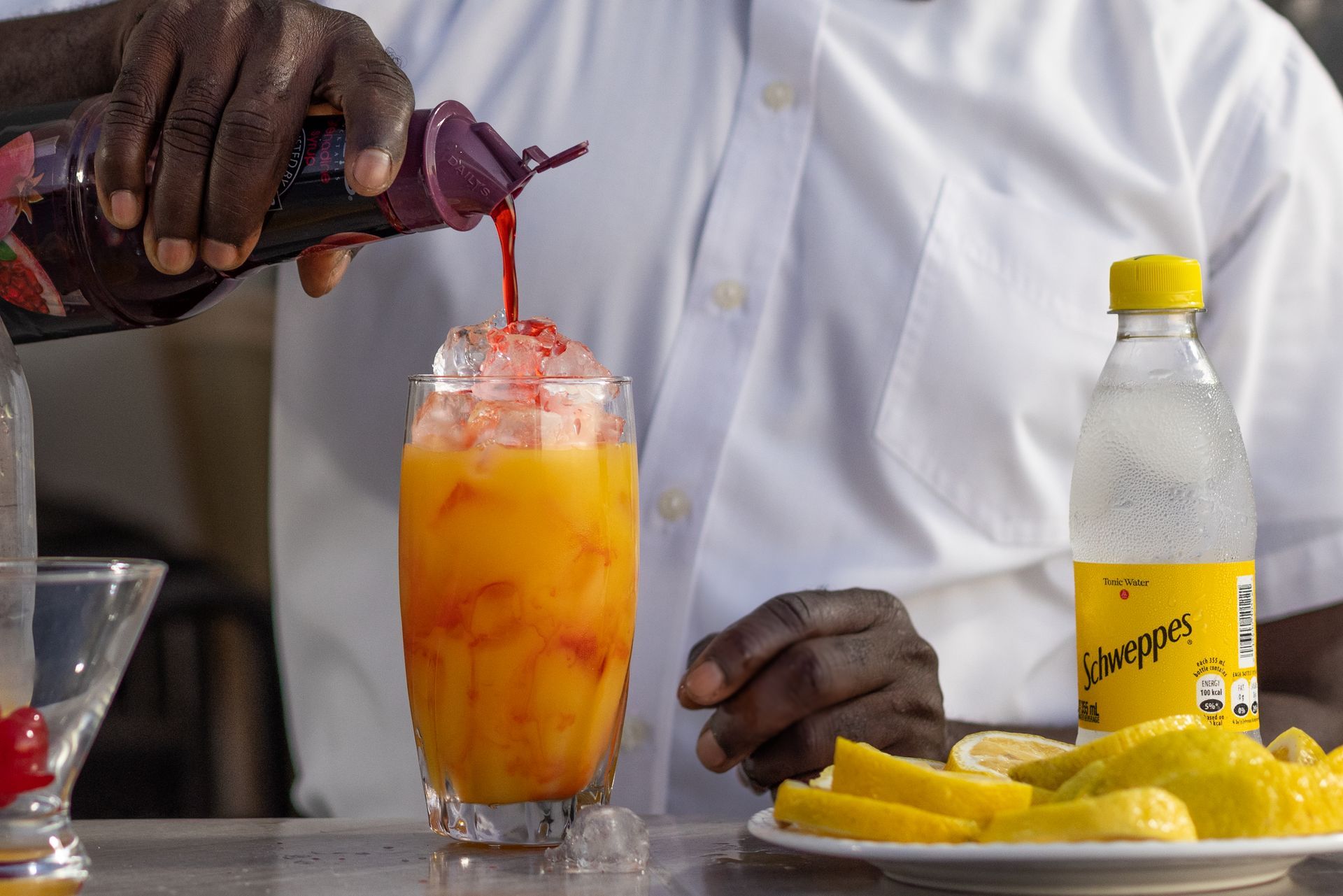 Bartender pouring red liquid into a tall glass of orange liquid with ice. A bottle of Schweppes and lemon slices are nearby.