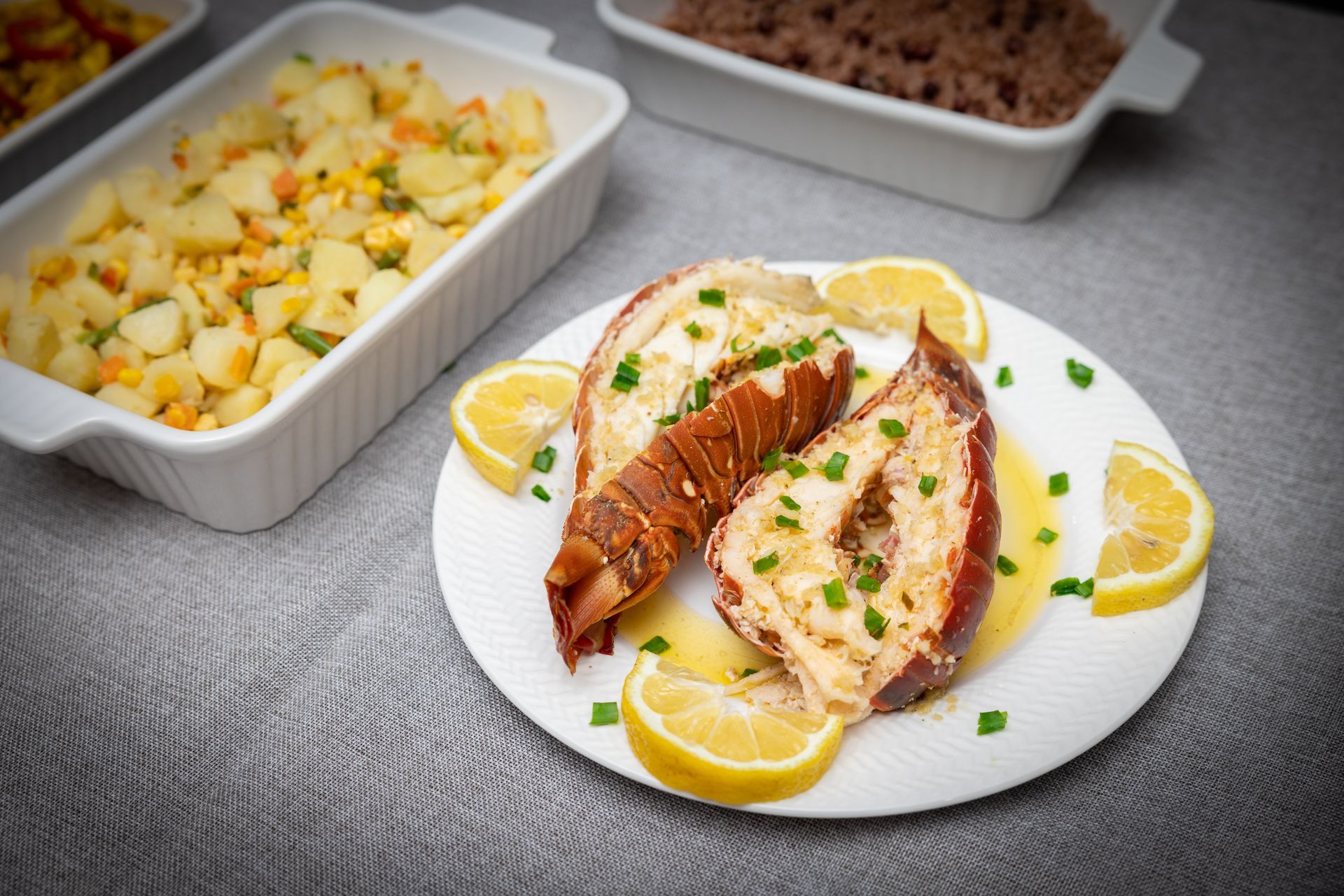 Lobster tail on a plate with lemon slices, side dishes of potato salad and meat.