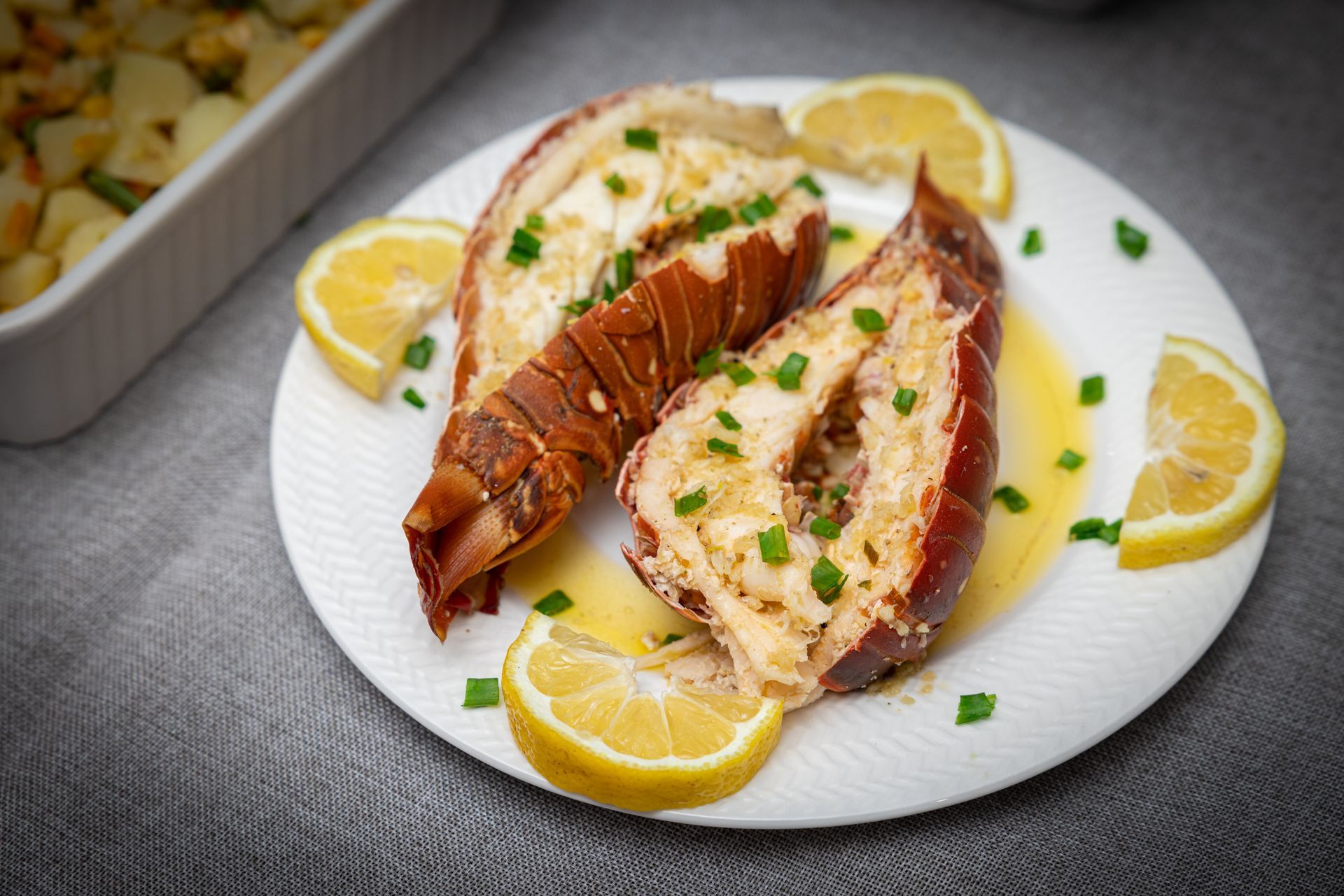 Grilled lobster on a white plate with lemon wedges and green chives; side dish in the background.