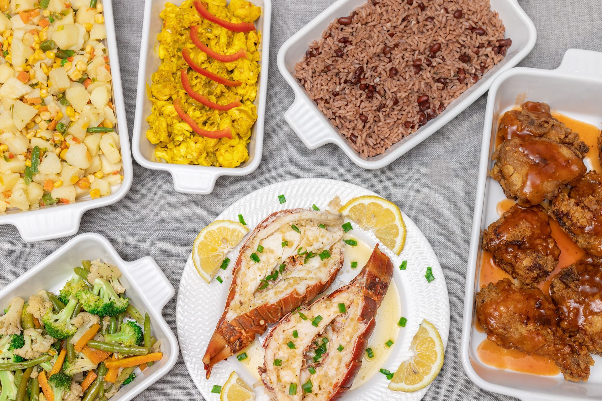Assortment of prepared dishes, including lobster, chicken, rice, vegetables, and potatoes, in white serving dishes.