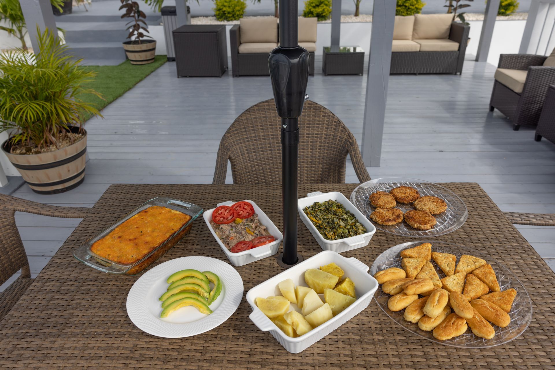 Outdoor dining table with dishes: casserole, avocado, vegetables, and snacks, surrounded by patio furniture.