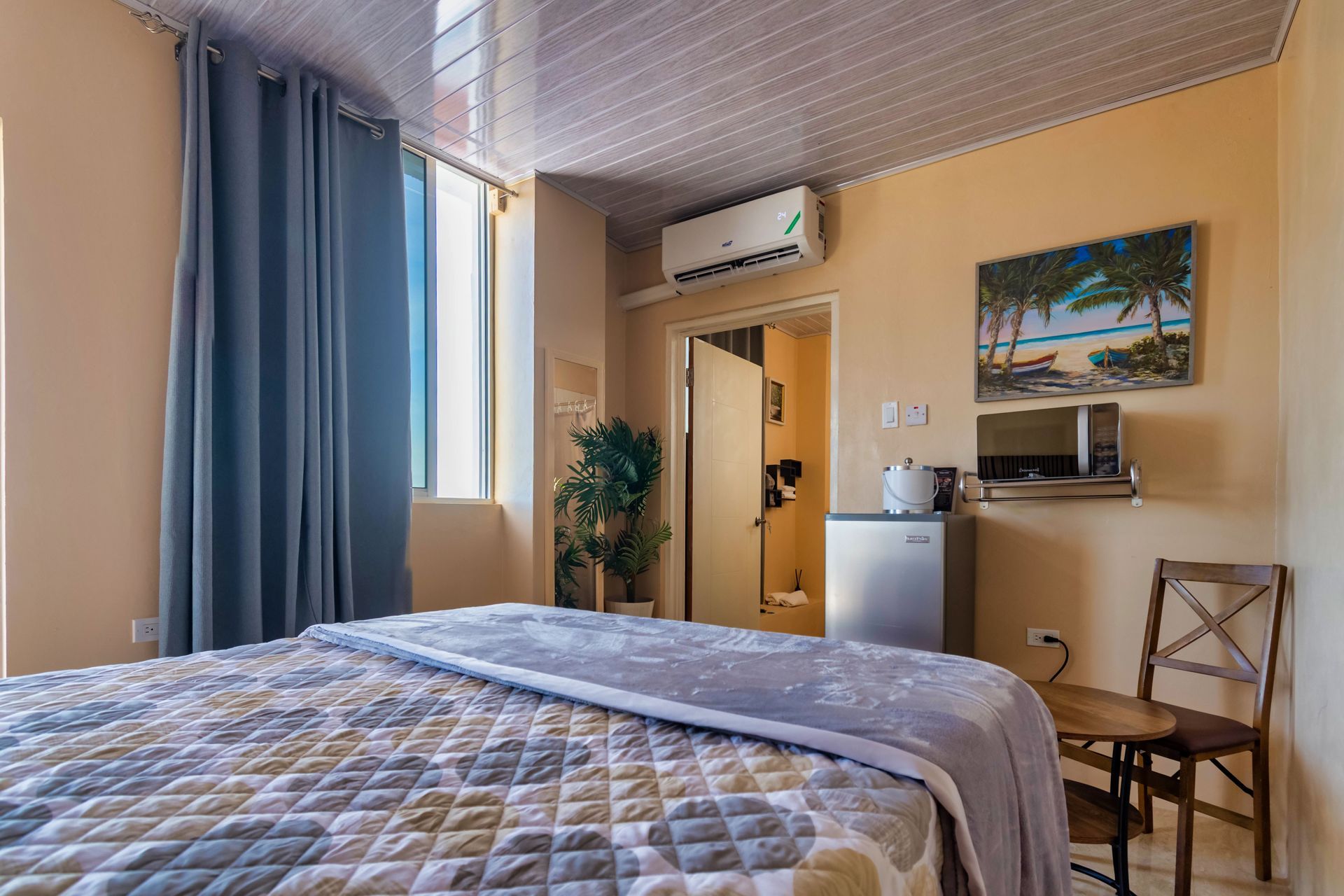 Bedroom with bed, blue curtains, small refrigerator, microwave, and beach art.