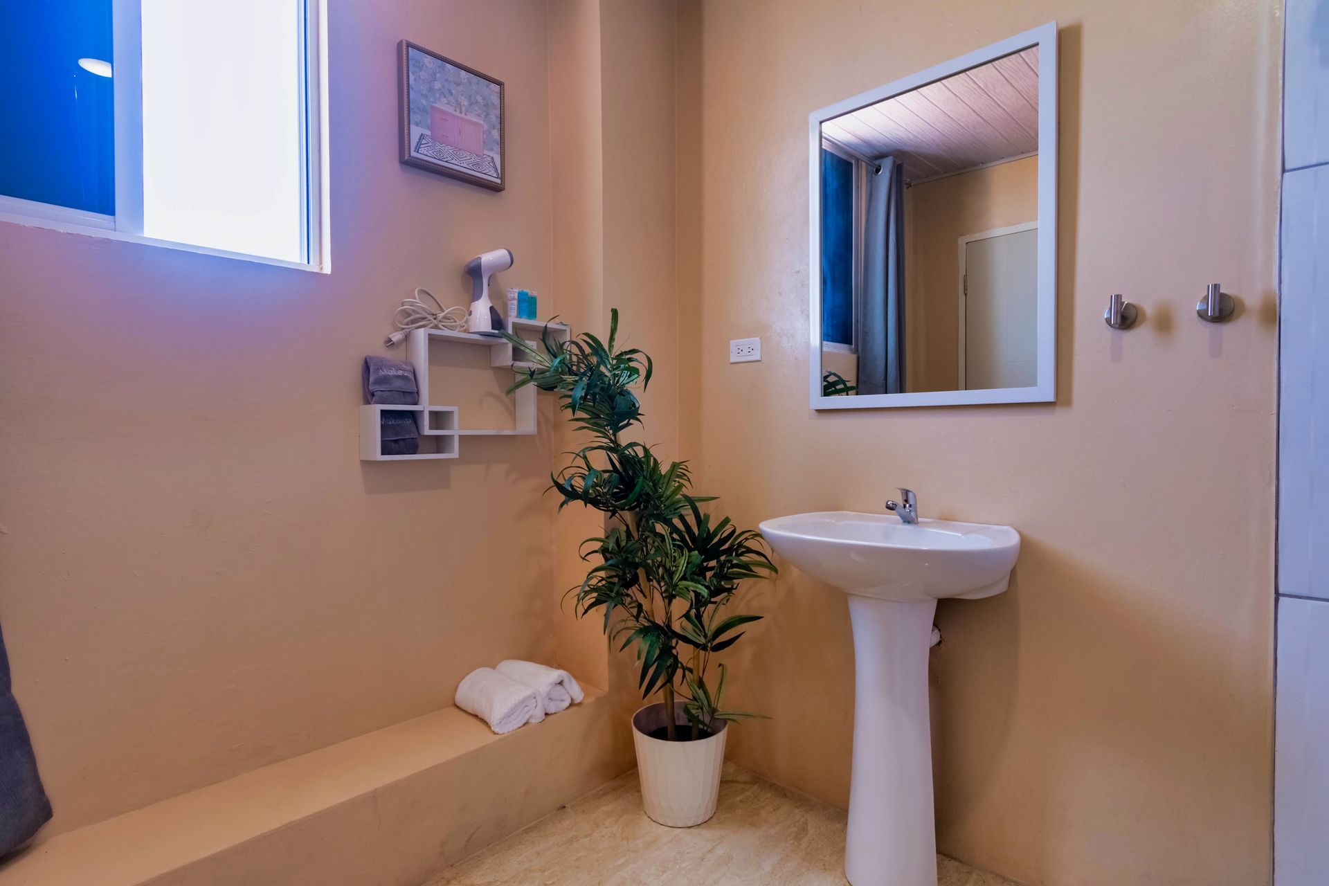Bathroom with a pedestal sink, mirror, small window, and a potted plant. Beige walls.
