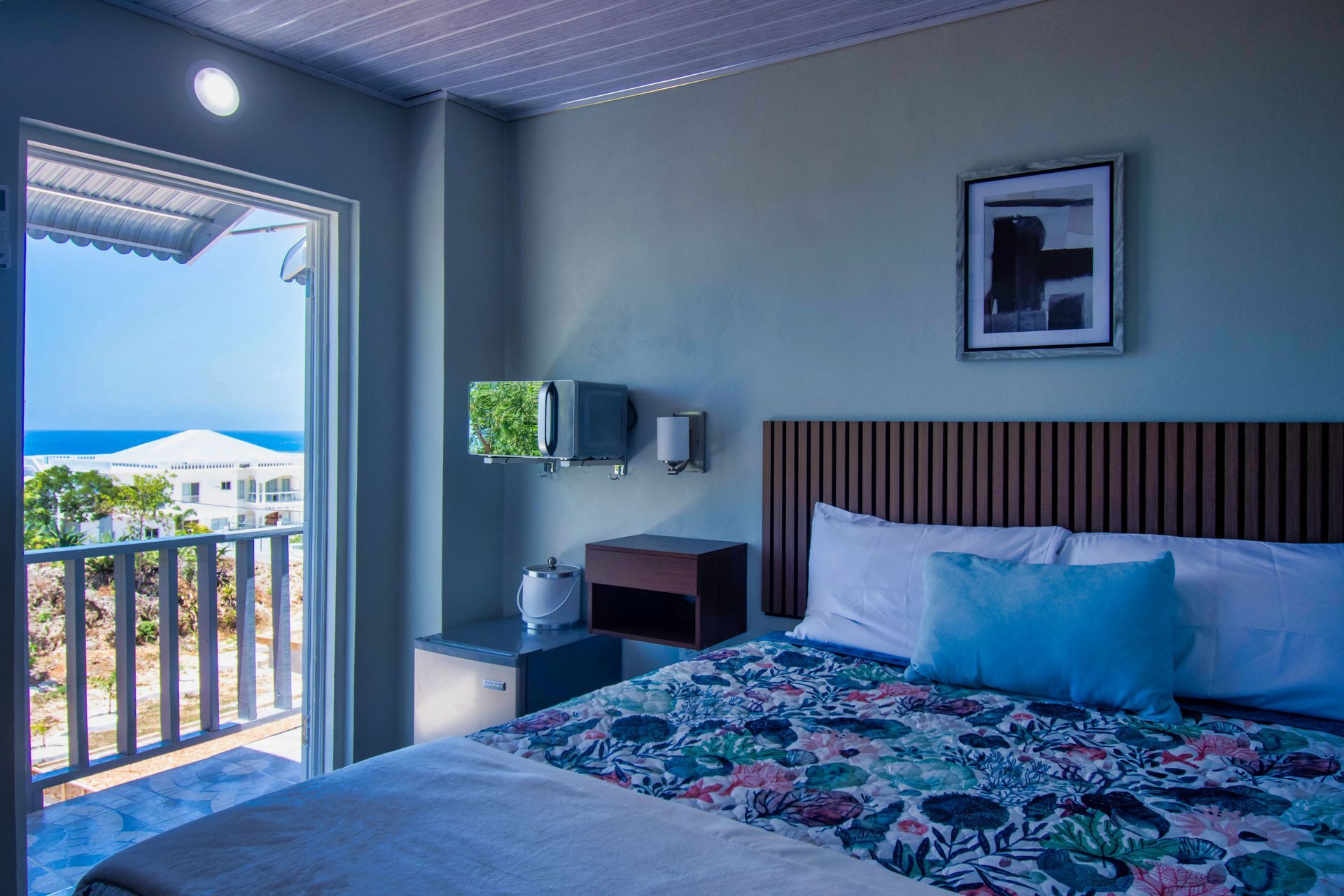 Bedroom with balcony overlooking the ocean. Light blue walls, floral bedspread, artwork, and a wooden headboard.