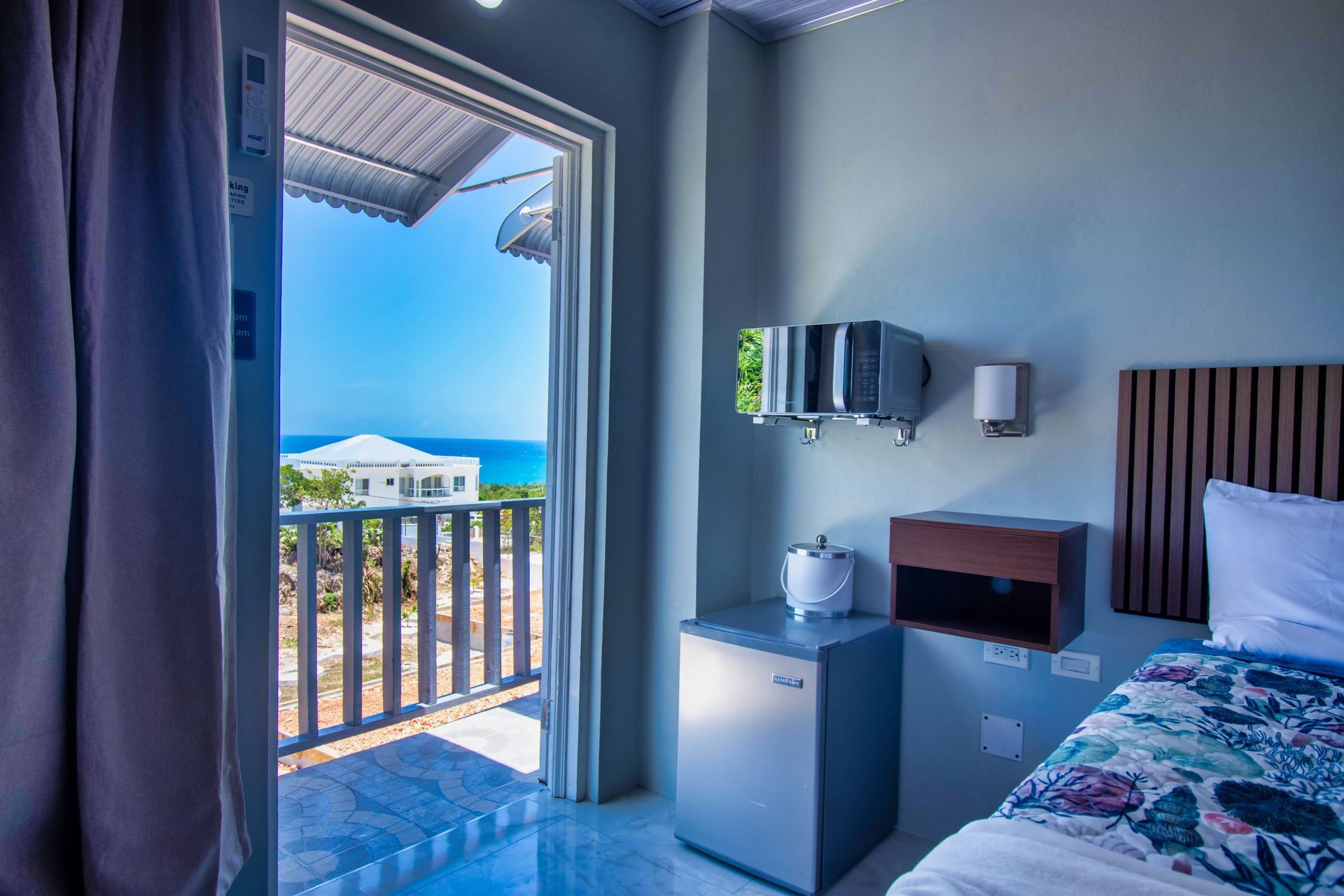 A bedroom with a balcony overlooking the ocean. Light blue walls, bed, and small refrigerator.