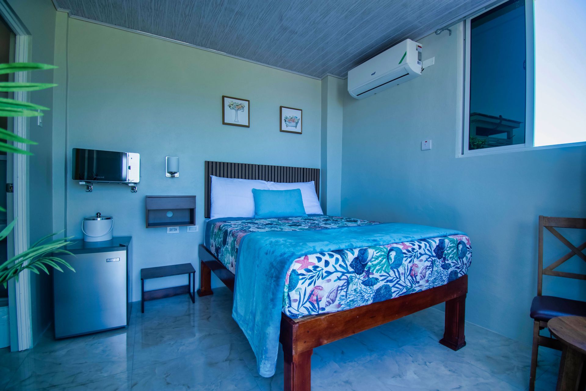 Bedroom with a bed, small fridge, and air conditioner; light blue walls and floral bedding.