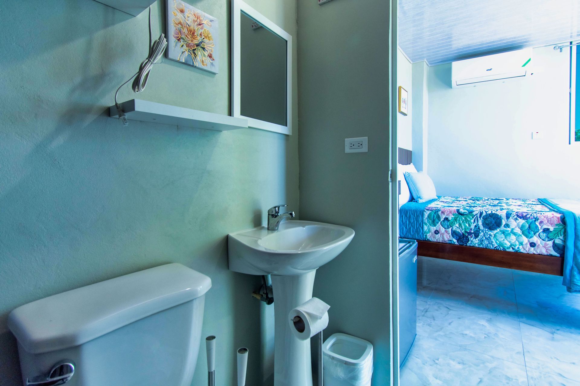 Bathroom with toilet, sink, mirror, and a doorway to a bedroom with bed and air conditioner.