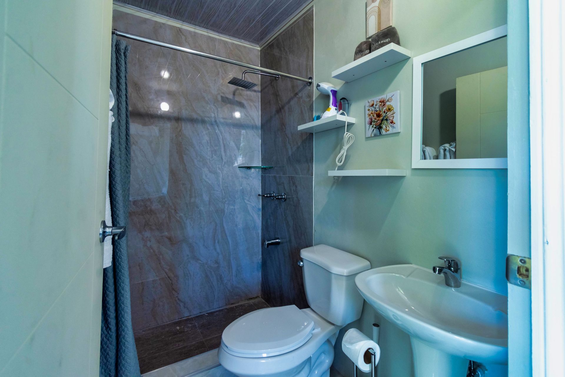 Bathroom with a toilet, sink, and shower. Gray walls and a light green wall, white shelves, and a mirror.