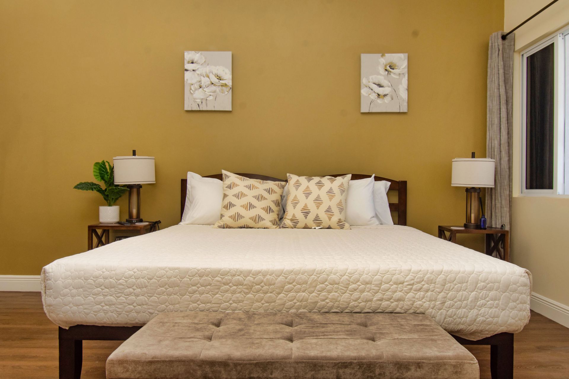 Bedroom with a bed, nightstands, lamps, and artwork against a yellow wall. A window is on the right.