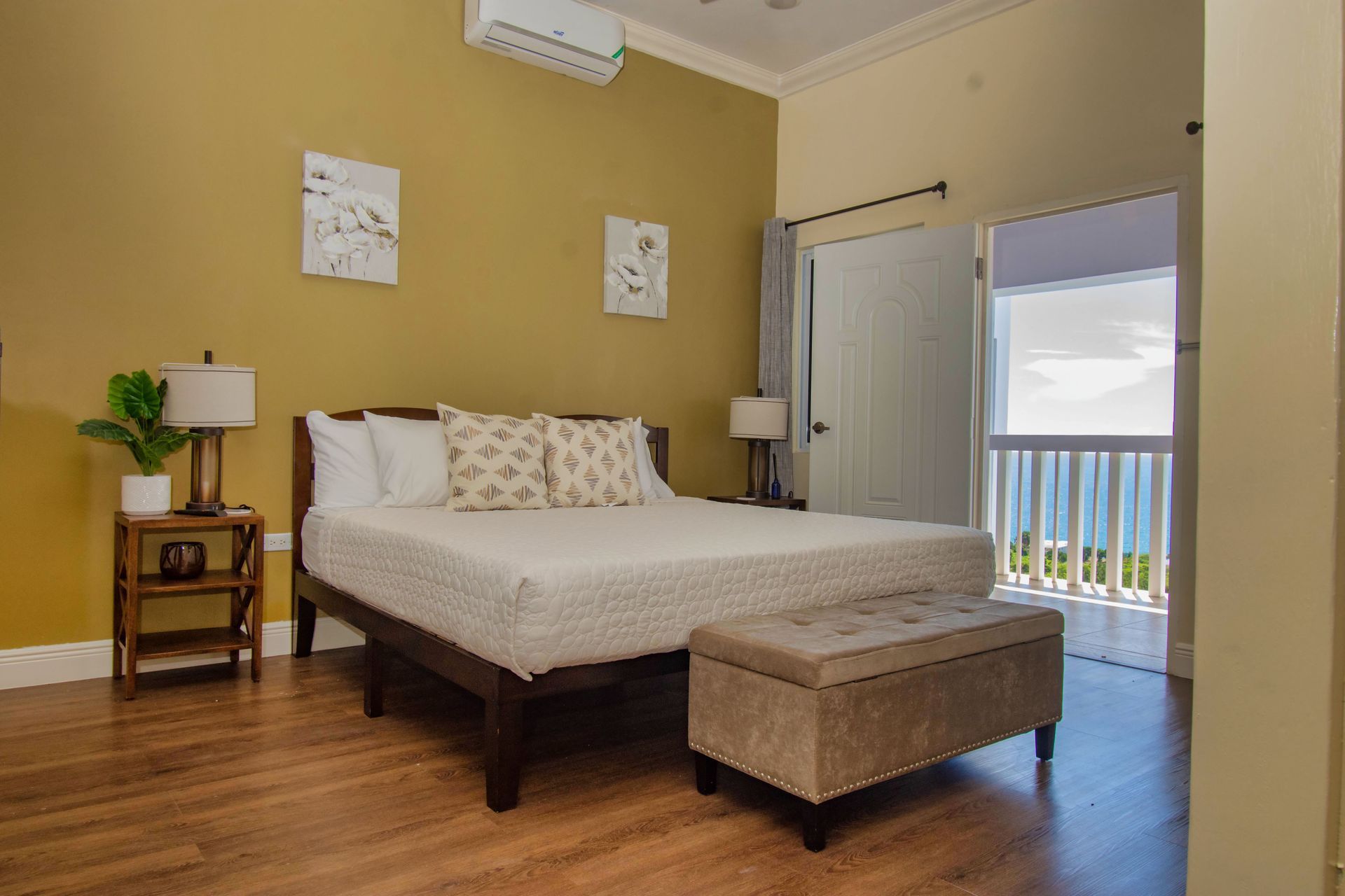 Bedroom with a bed, bench, and open door to a balcony overlooking the ocean. Green walls, wooden floors, and soft lighting.