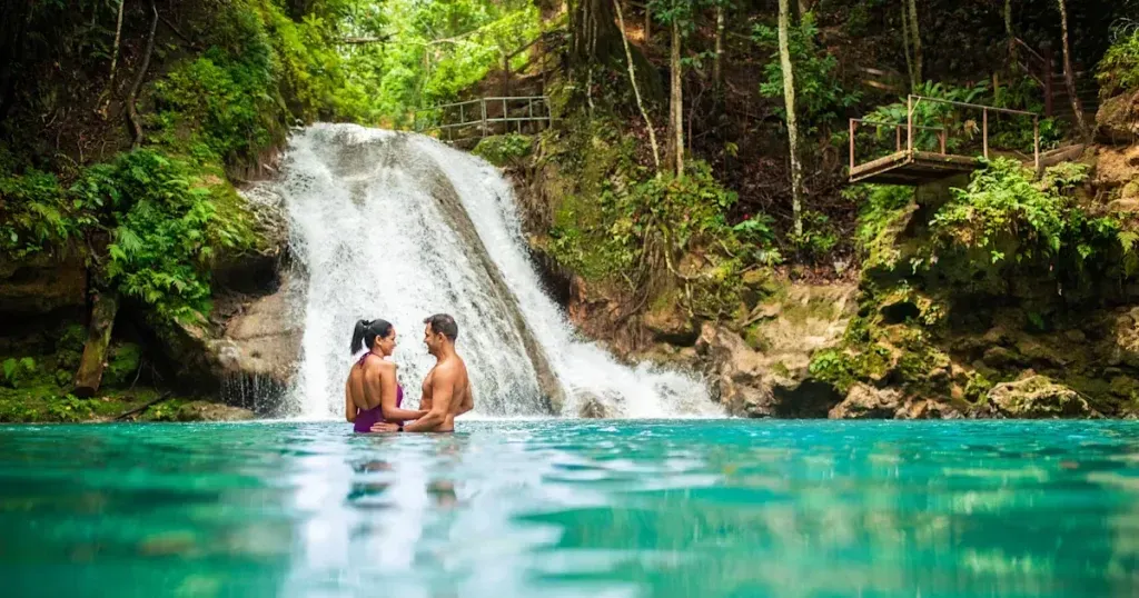 Couple in turquoise water, under a waterfall, surrounded by lush green foliage.