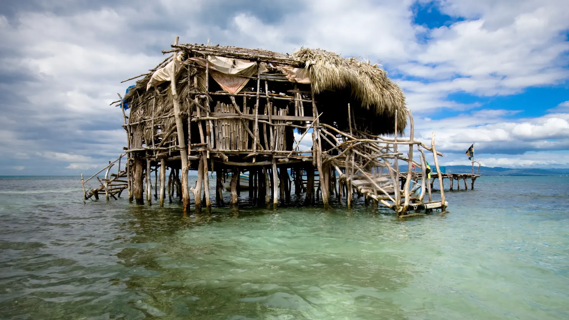 Stilt-house in ocean with weathered wooden structure and thatched roof under a partly cloudy sky.