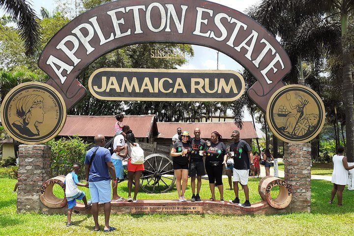 Appleton Estate Jamaica Rum sign with people in front. Brown archway over entryway. Green grass and trees visible.