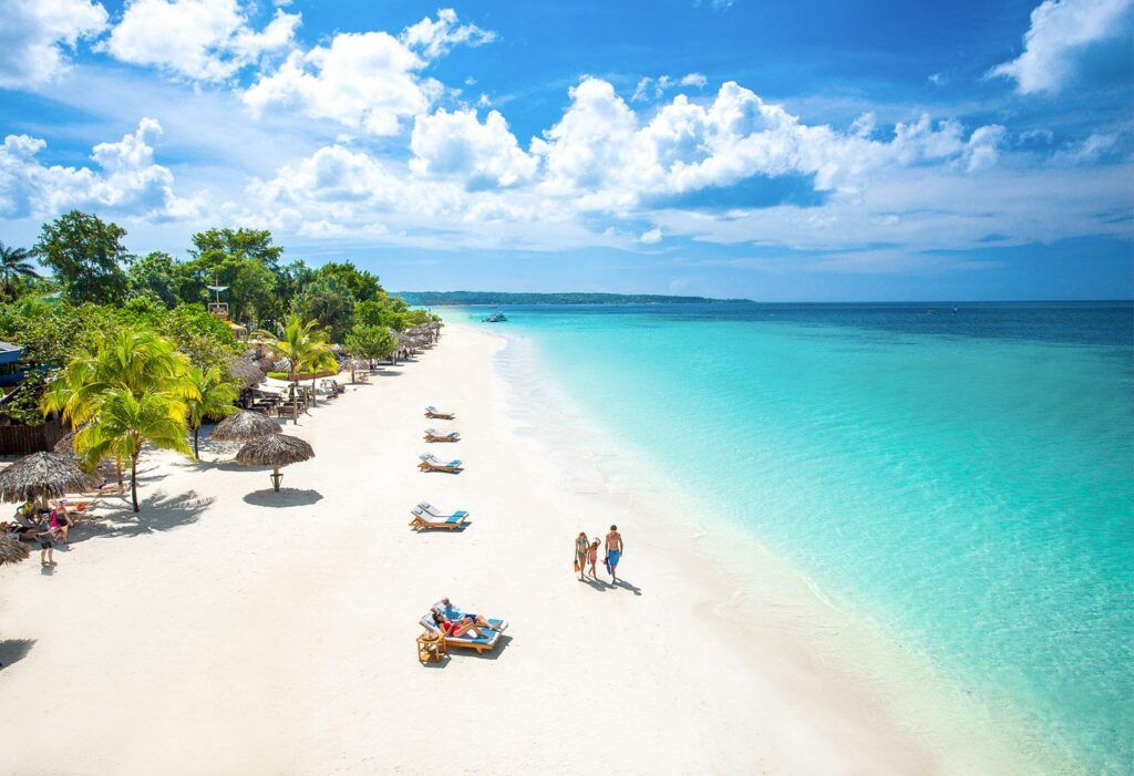 White sand beach with turquoise water, palm trees, thatched umbrellas, and people walking.