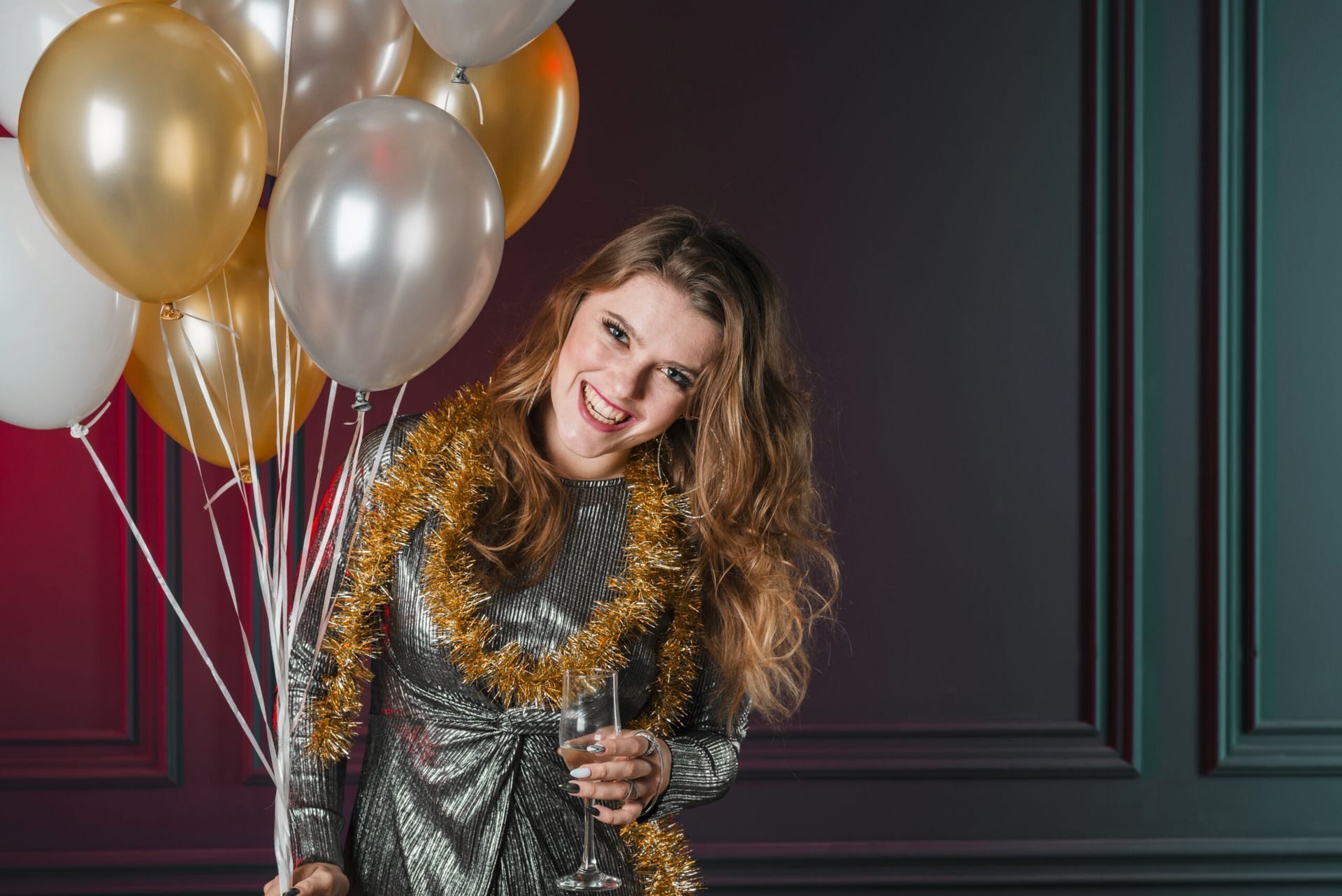 Woman in a silver dress with gold tinsel, holding balloons and a champagne glass, smiling.