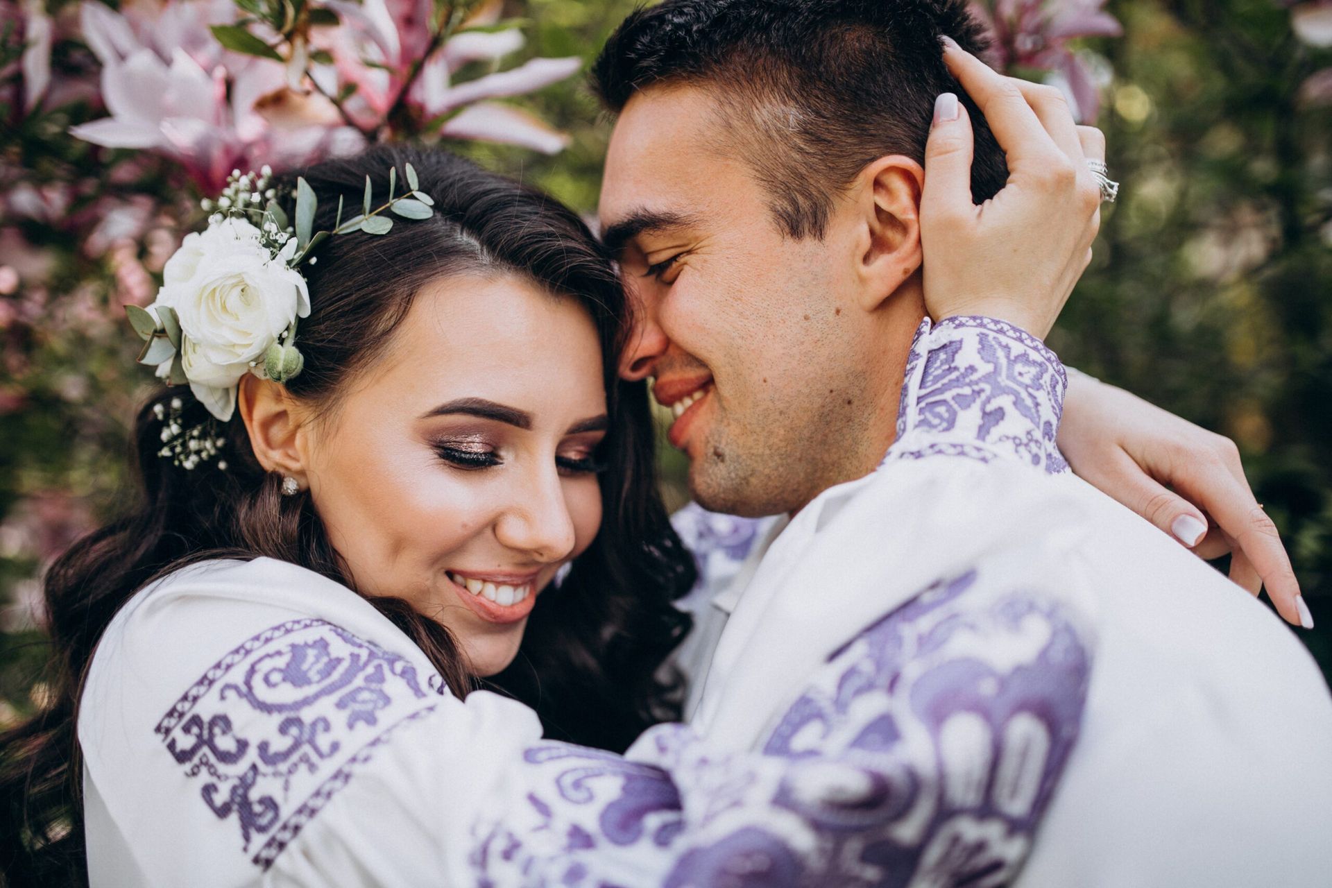 Smiling couple embraces outdoors, near blooming pink flowers, wearing white shirts with purple embroidery.