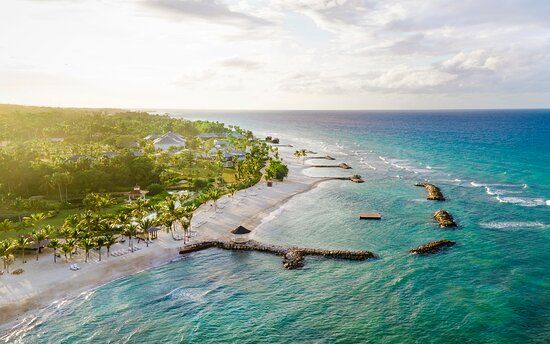 Aerial view of a tropical beach with white sand, turquoise water, and lush green vegetation under a sunny sky.