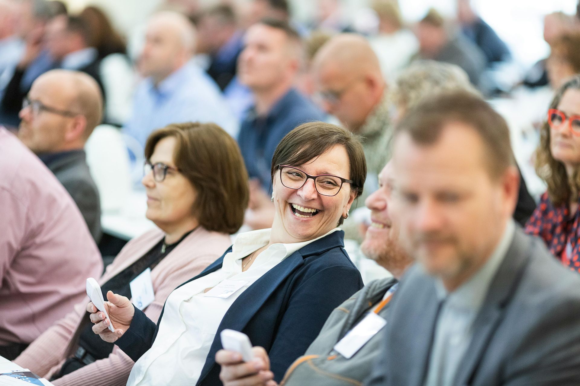 People at a conference; woman in glasses laughs, holding a device. Others in background.