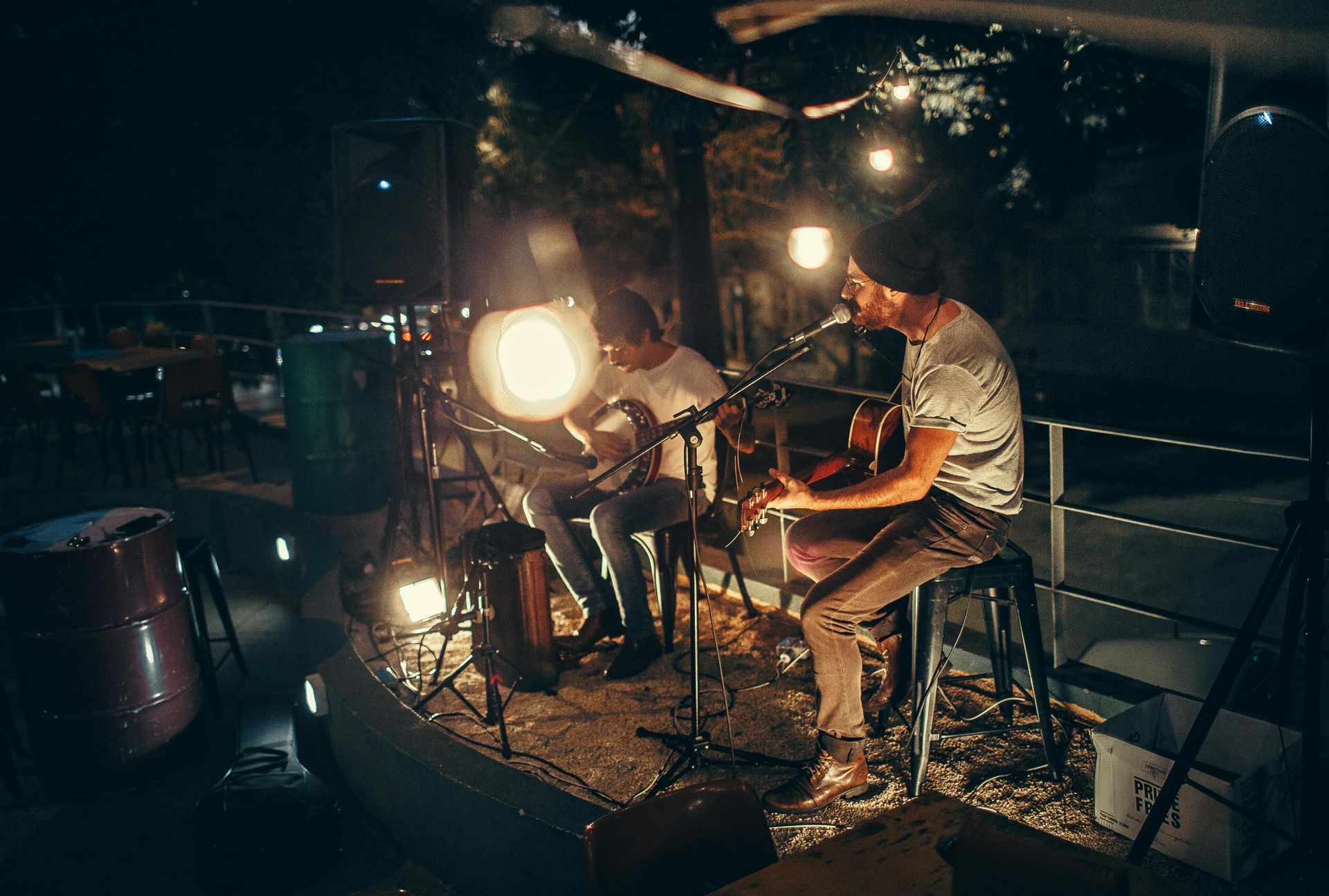 Two musicians playing acoustic guitars under warm string lights at night.