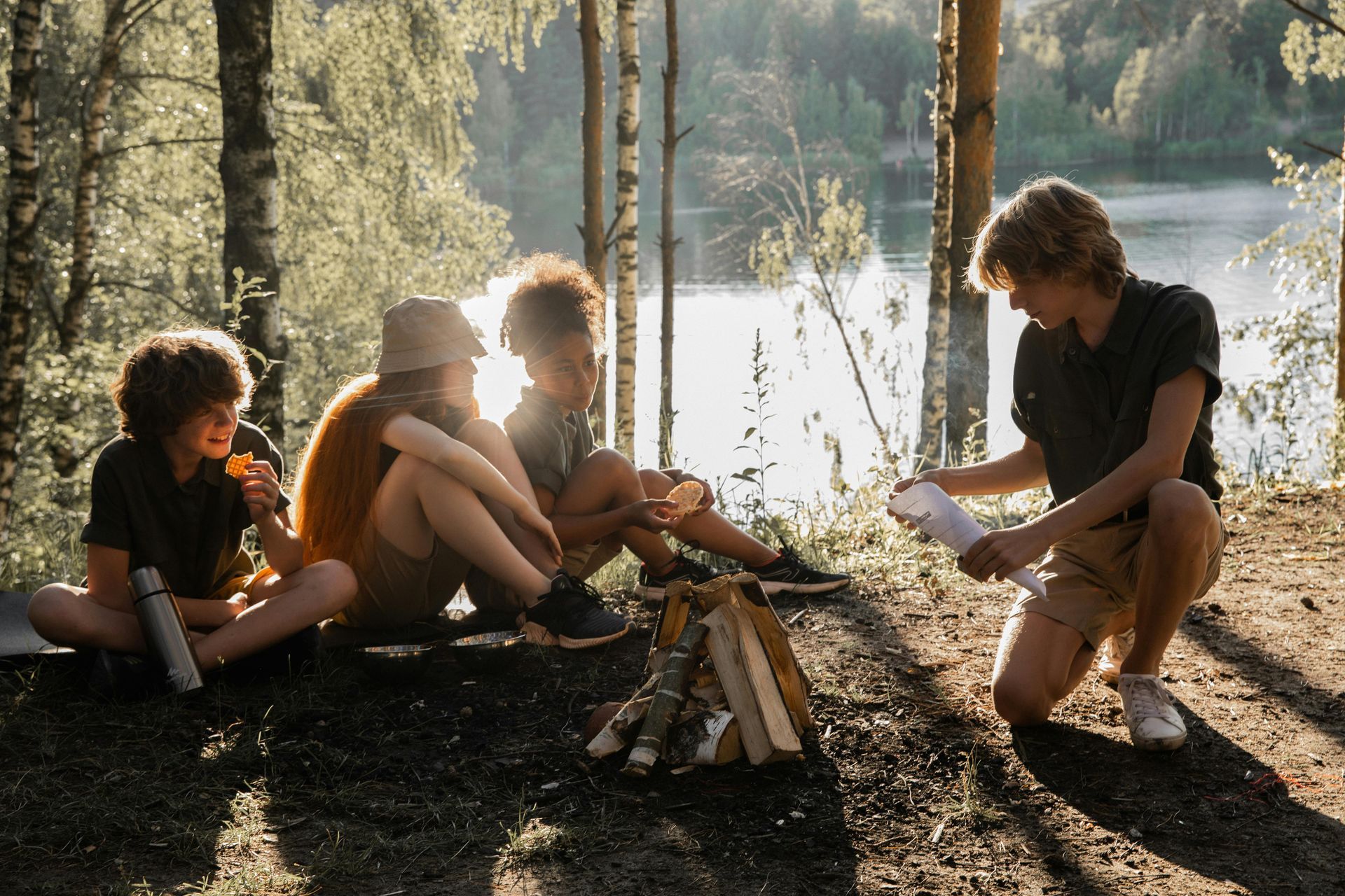 Friends gather around a small campfire by a lake, reading a map in a sunlit forest.