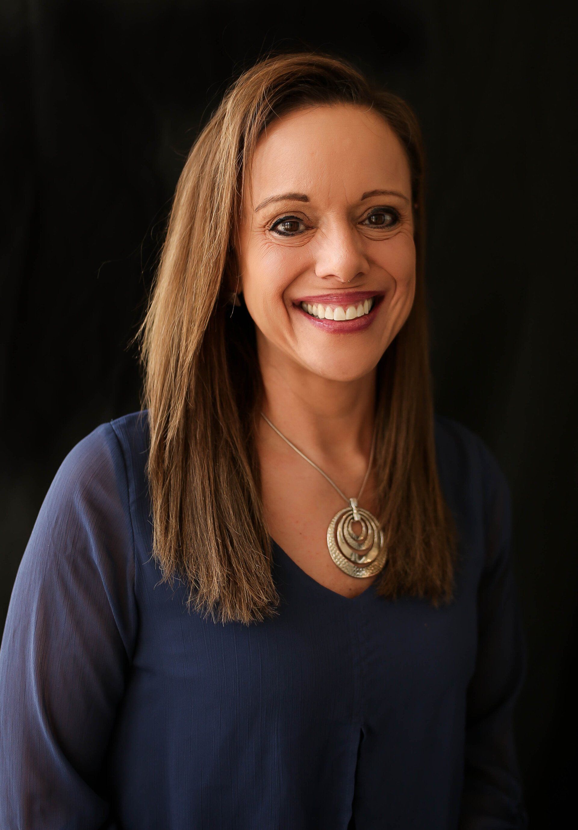 A woman wearing a blue shirt and a necklace is smiling for the camera.