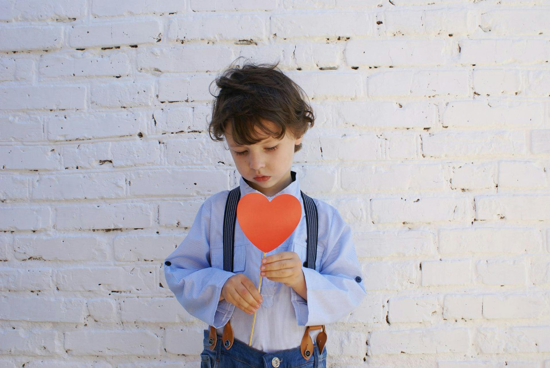 A young boy is holding a red heart in front of a white brick wall.