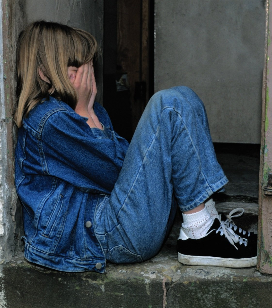A girl in a denim jacket sits on the steps covering her face