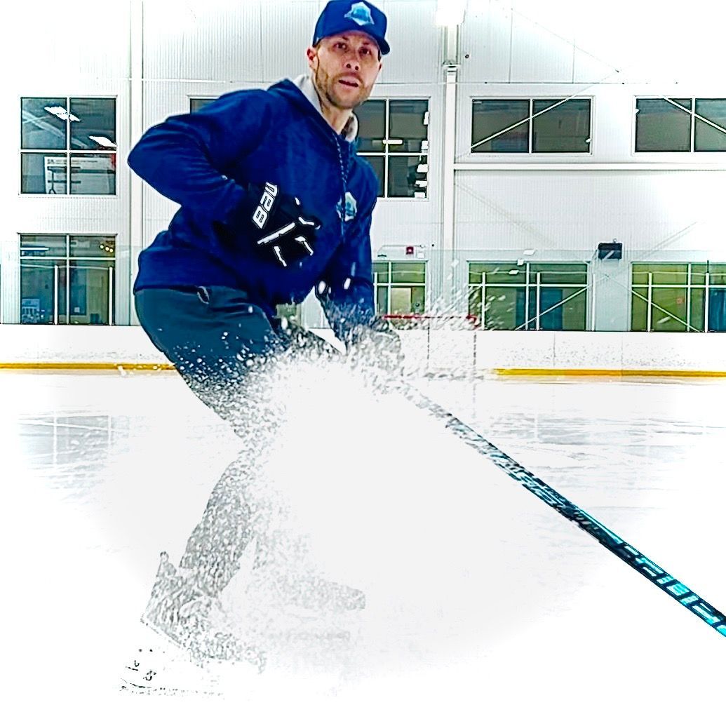 A person in a blue hoodie and cap performs a hockey stop on an ice rink, creating a large spray of ice.