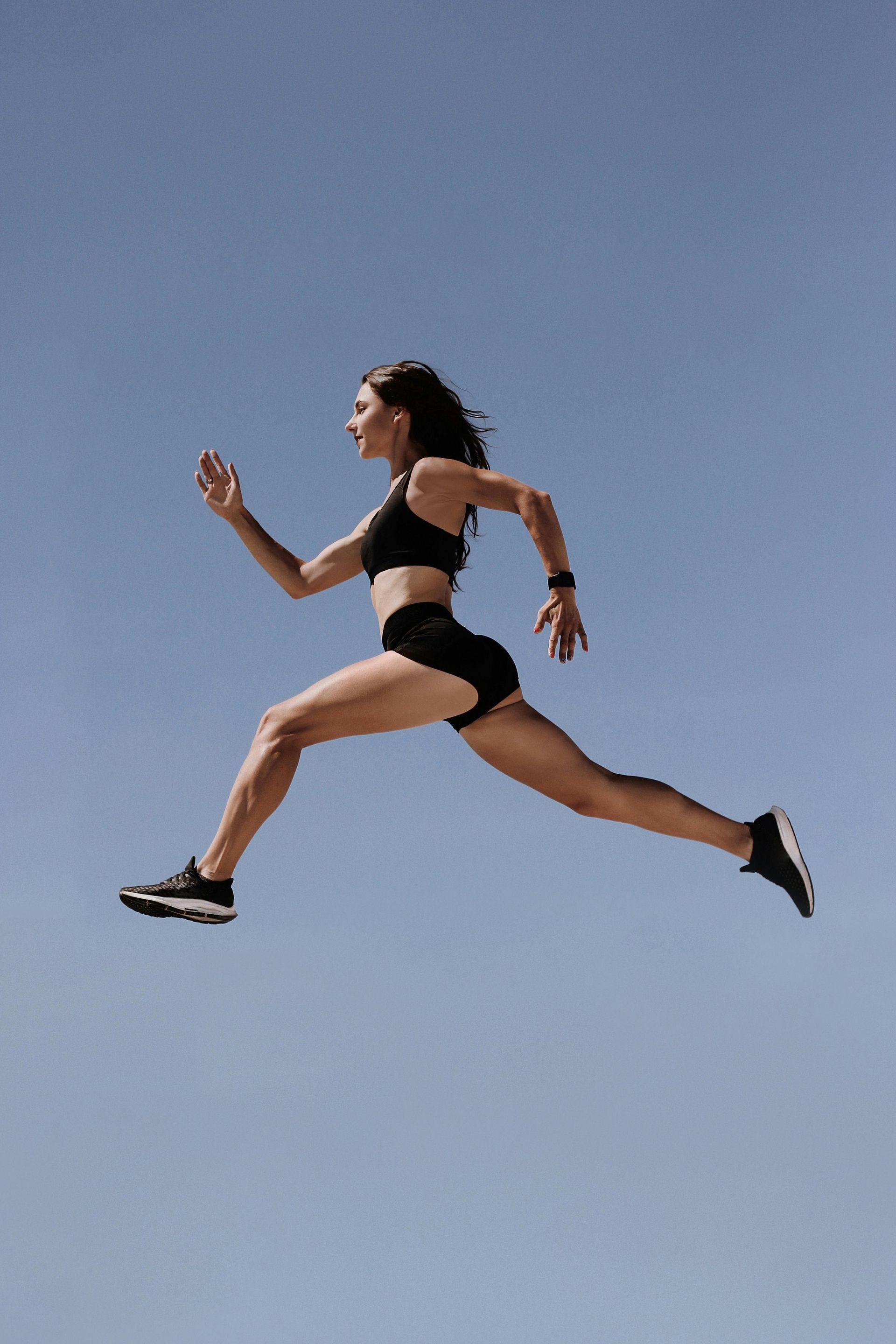 Woman mid-air, running in black athletic wear against a blue sky.