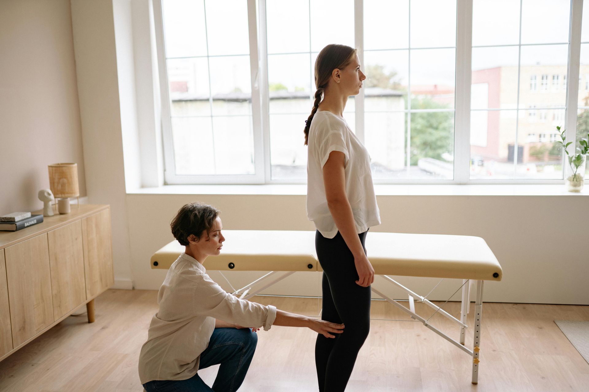 Woman receiving physical therapy; therapist checking leg position. Bright room, massage table.