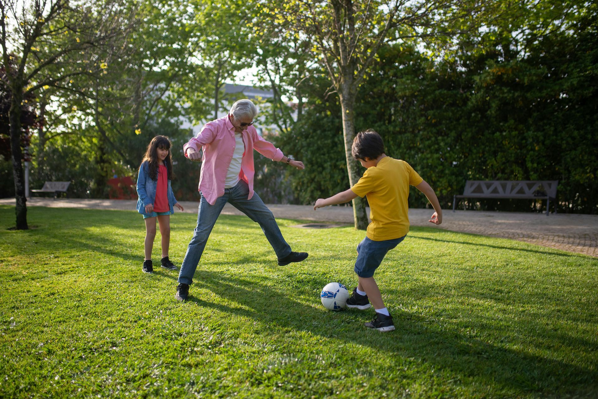 Man and boy playing soccer in a park, girl watches. Green grass, sunny day.