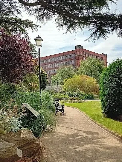 Pathway through a garden with a red brick building in the background. A bench and lamppost are visible.
