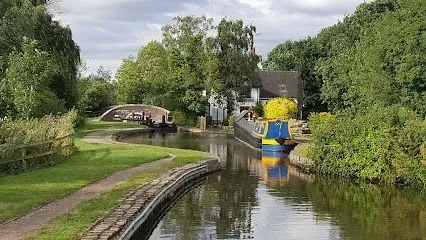 Canal scene with narrow boat at lock, arched bridge, pathway, trees, and house.