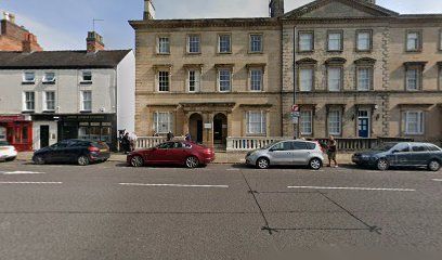 Row of buildings with parked cars in front; people walking on the sidewalk.