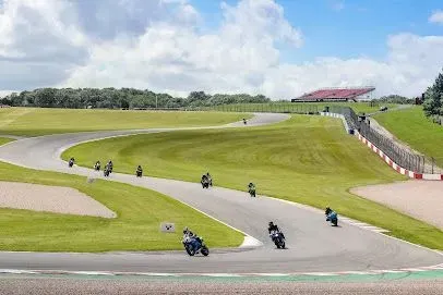 Motorcycles racing on a curved track, sunny day, green grass, spectator seating in the background.