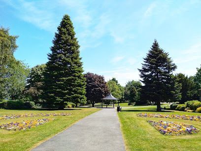 Pathway through a park with tall trees, flower beds, and a gazebo under a bright blue sky.