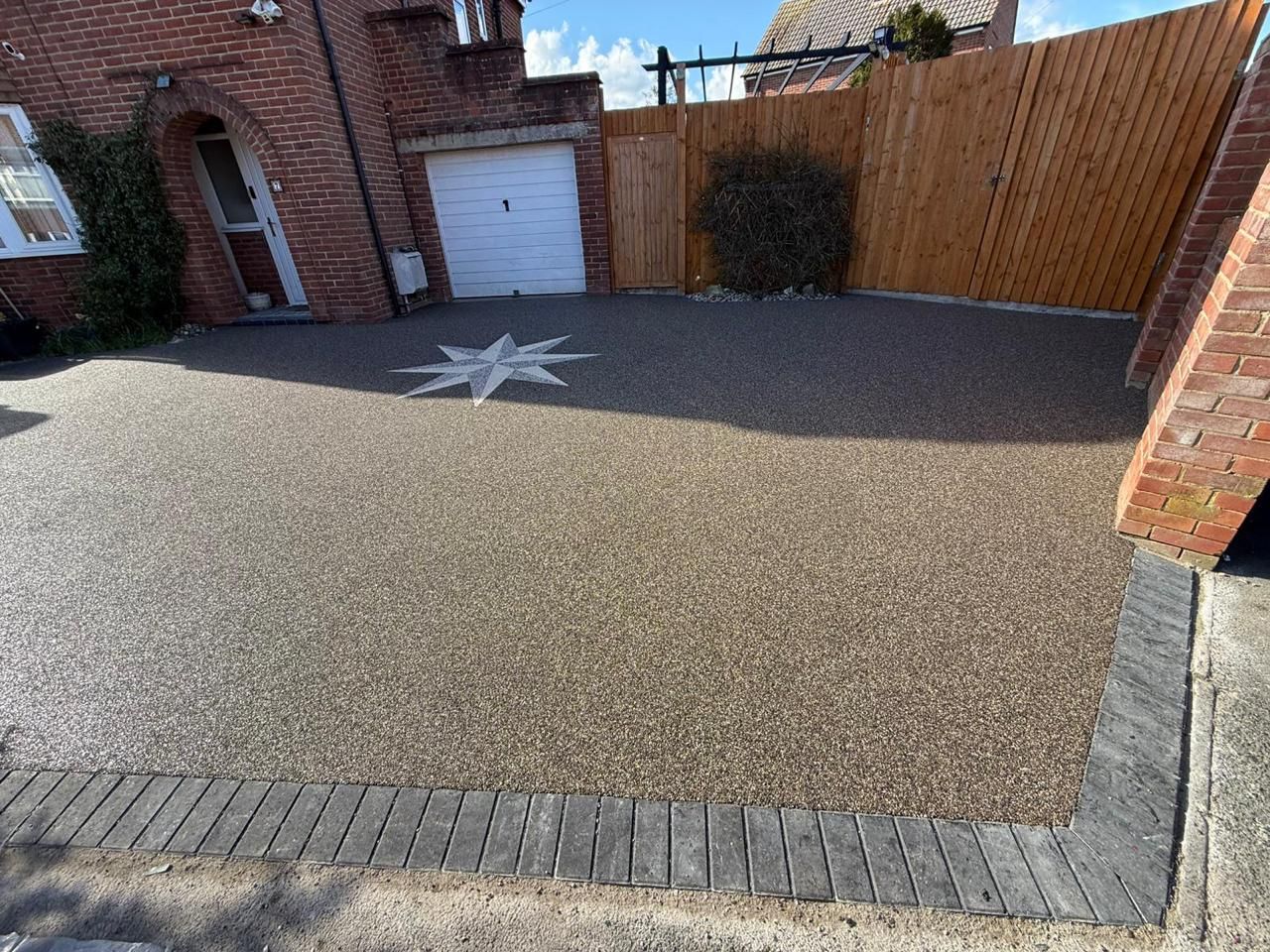 A resin-bound driveway featuring a white compass rose design, edged with dark grey block paving near a brick house.