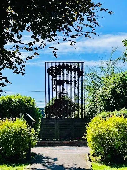 Monument of a man's face framed by metal grid, surrounded by green foliage, blue sky background.