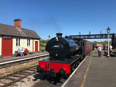 Black steam train at a station. Red and white buildings, people waiting. Clear, blue sky.